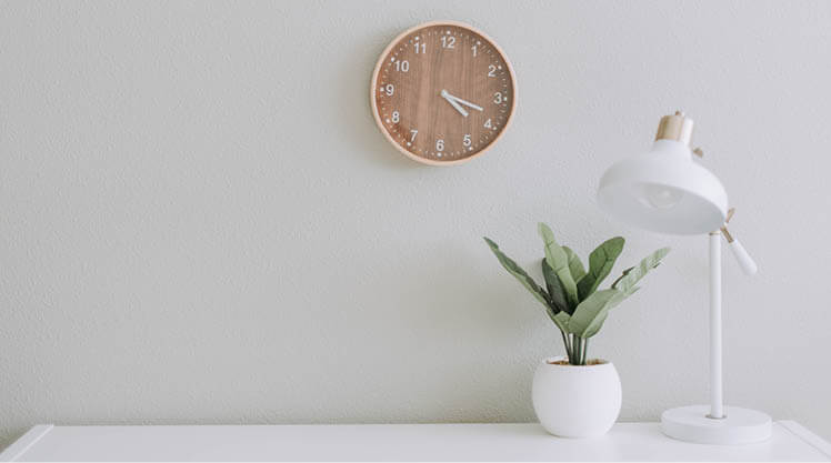 A white lamp and green plant sit on a desk, a wall clock behind them.